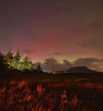 A beautiful display of the Aurora Borealis illuminating the night sky over a tranquil lake in Connemara, County Galway, with mountains silhouetted in the background.